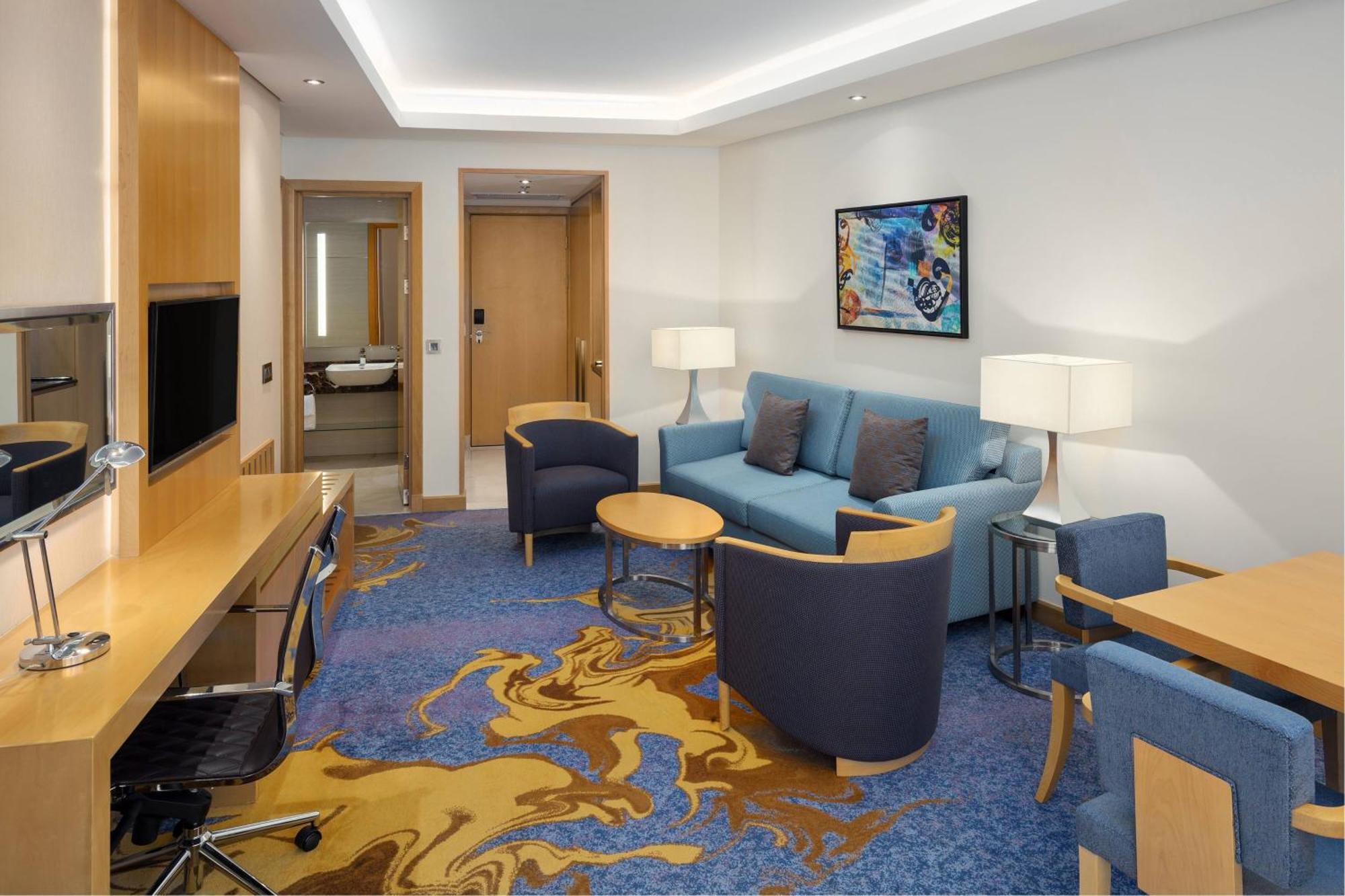 Sheraton Makkah Jabal Al Kaaba Hotel Мекка Экстерьер фото The photo displays a modern hotel room or suite. The room features a comfortable seating area with a blue couch and two blue armchairs surrounding a small round table. A wooden desk with a black office chair is positioned against one wall, accompanie