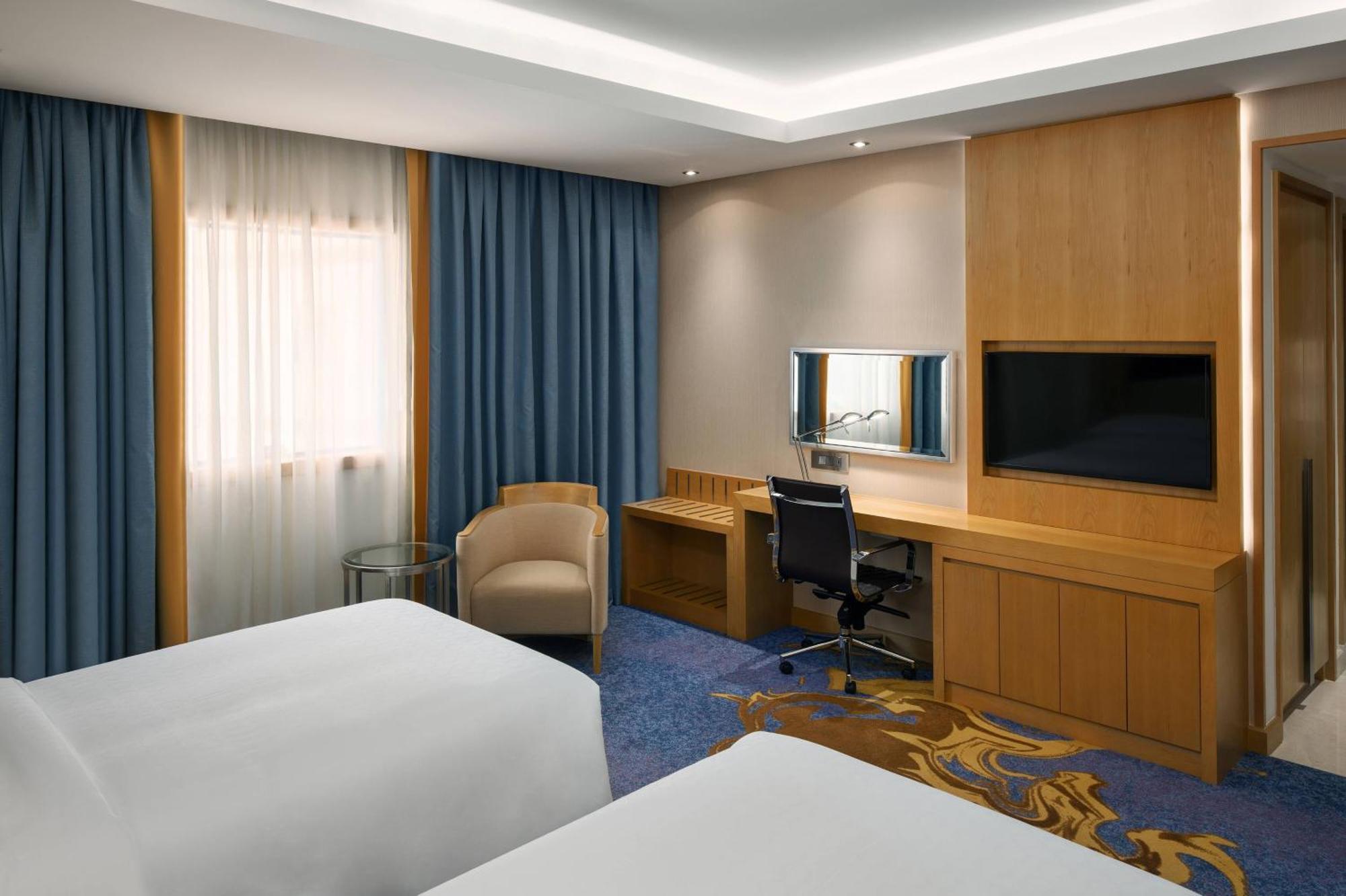 Sheraton Makkah Jabal Al Kaaba Hotel Мекка Экстерьер фото The photo shows a hotel room featuring two beds with white linens. There are large windows covered with blue curtains that allow natural light to enter. On one side of the room, there is a wooden workstation with a chair and a mirror, along with a fl