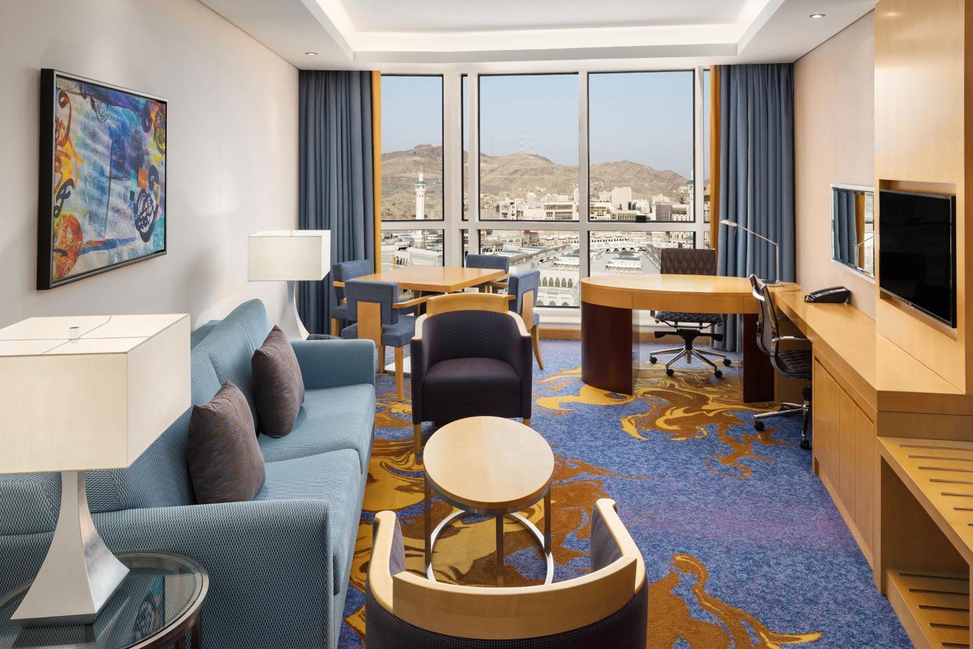 Sheraton Makkah Jabal Al Kaaba Hotel Мекка Экстерьер фото The photo shows a well-decorated hotel room featuring a modern and comfortable interior. There is a cozy seating area with a blue sofa and armchairs, complemented by a round coffee table. A desk with a chair is also present, ideal for work. The room 