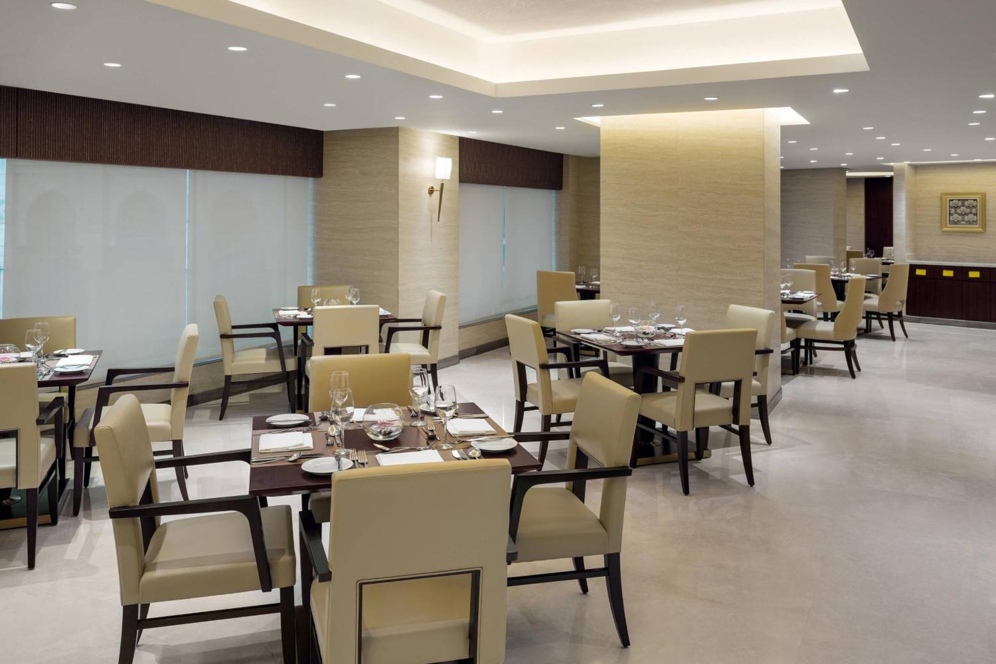 Sheraton Makkah Jabal Al Kaaba Hotel Мекка Экстерьер фото The photo shows a modern restaurant interior. It features several dining tables, each set with utensils and tableware. The chairs are upholstered in a light fabric, and the overall decor is sleek and minimalistic, with soft lighting from overhead fix