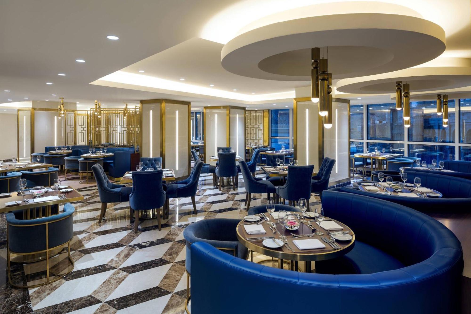 Sheraton Makkah Jabal Al Kaaba Hotel Мекка Экстерьер фото The photo shows the interior of a modern restaurant. The design features elegant curved blue seating arranged around round tables with plates and glassware set for dining. The flooring is a striking marble pattern, adding to the upscale ambiance. The
